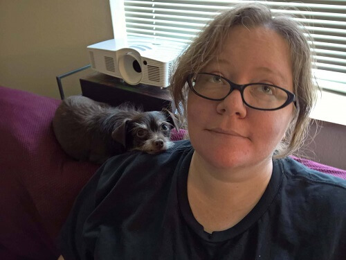 A photo of a woman wearing glasses, sitting on a couch, with a small dog resting his head on her shoulder.